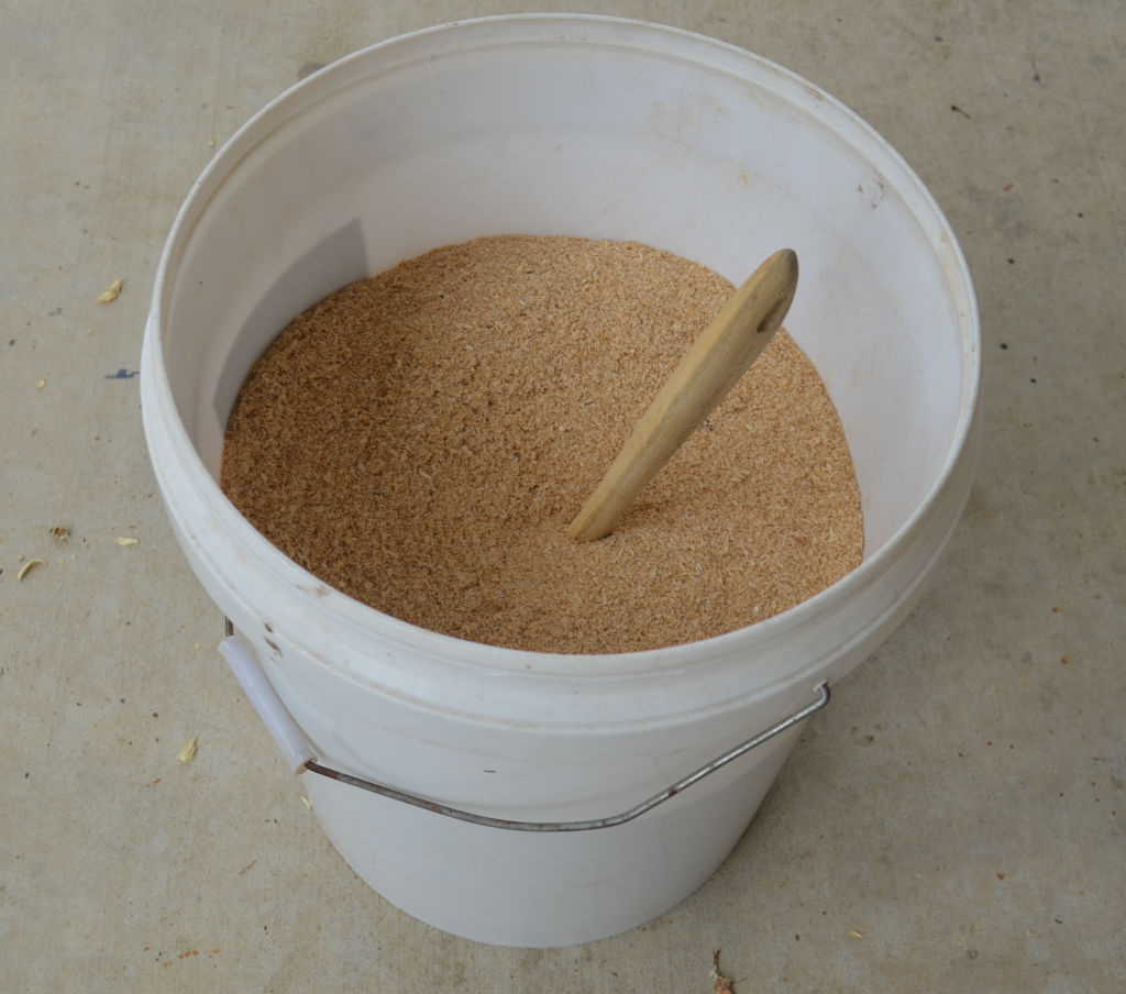 photo of a 20 liter container with bran and a wood spoon in it. The bran containers mealworm eggs which are mixed using the wood spoon using a method called average levelling.
