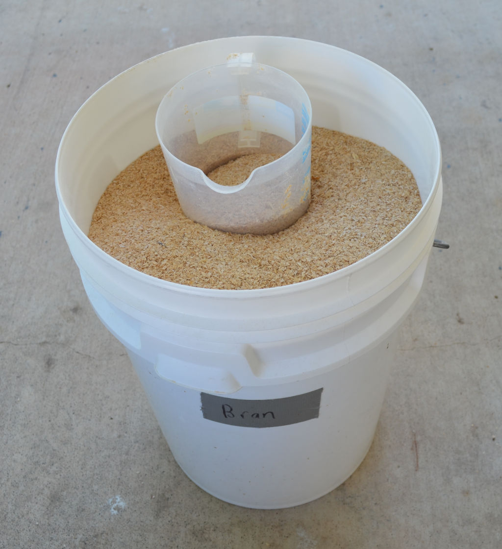 Photo of a 20 liter white bucket of wheat bran for commercial mealworm breeding. A measuring jug is at the top of the wheat bran to measure out bran.